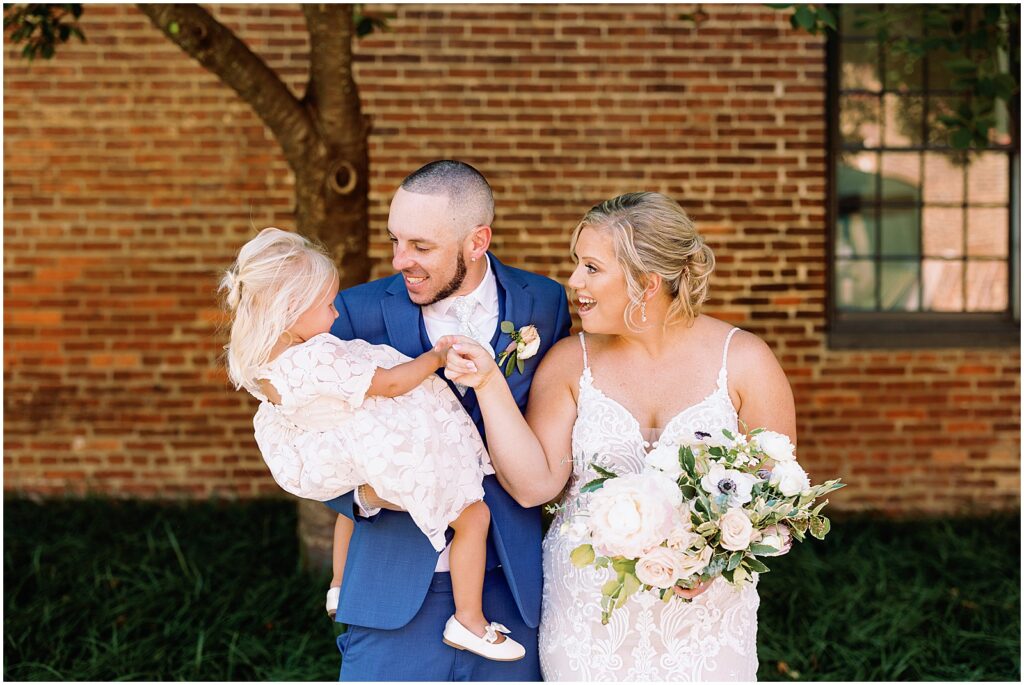 A bride and groom laugh while they hold a flower girl.