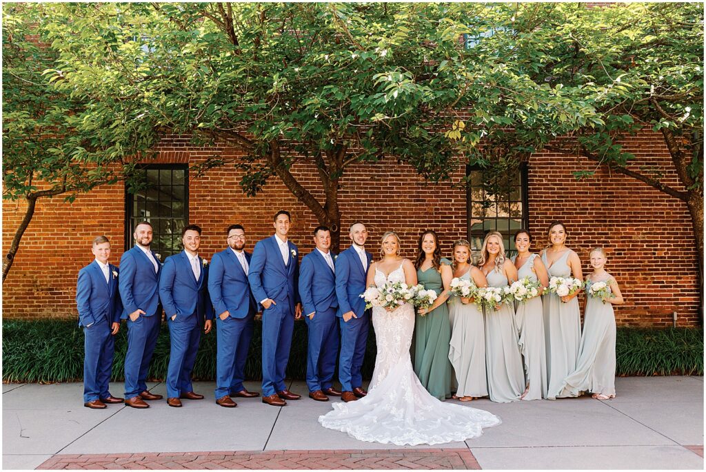 A bride and groom pose with their wedding party in blue groomsmen suits and green bridesmaids dresses.