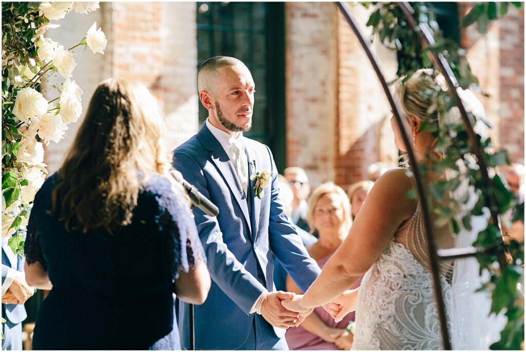 A groom holds a bride's hands and listens to a wedding officiant.