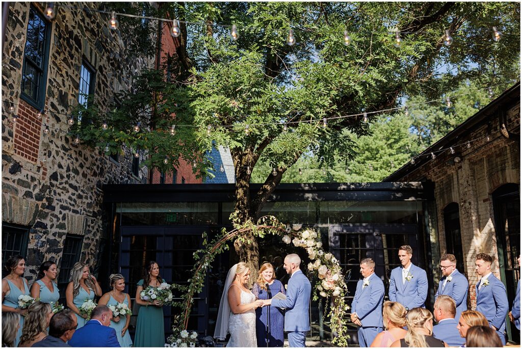 A wedding party stands on either side of a floral arch during a courtyard wedding ceremony.