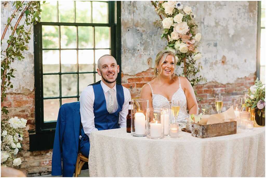A bride and groom smile at their sweetheart table while they listen to a wedding toast at Mt Washington Mill Dye House.
