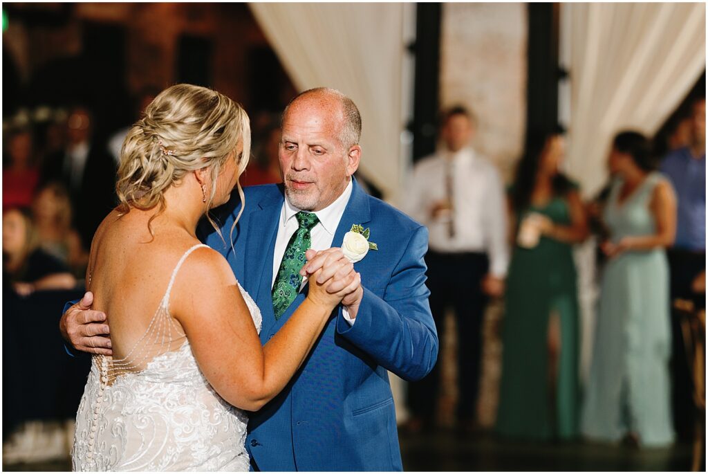 A bride dances with her father while wedding guests watch from the edge of the dance floor.