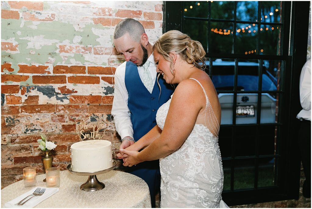 A bride and groom cut a wedding cake beside a window.