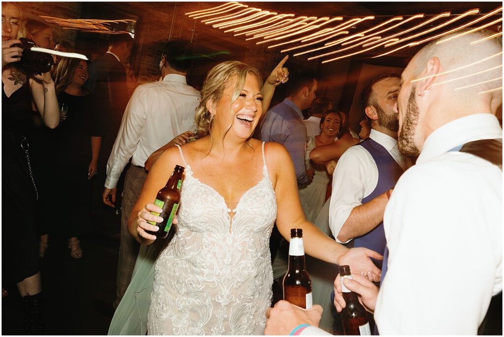 A bride laughs and carries a beer across the dance floor.
