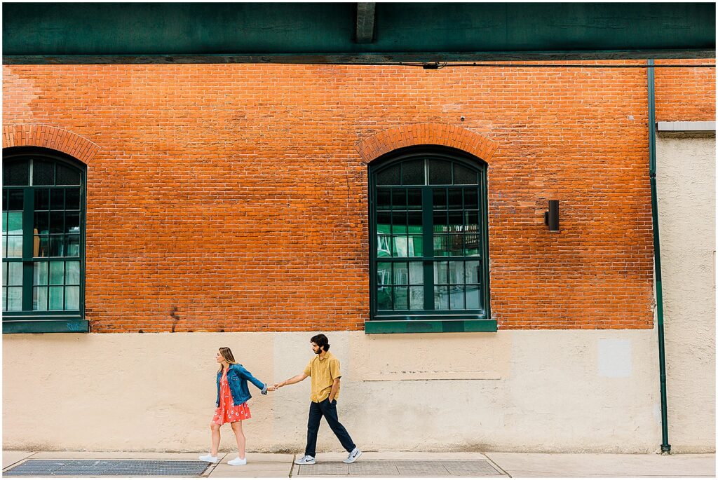 Brittany leads Joey past a brick building in a candid engagement photo in Fishtown.