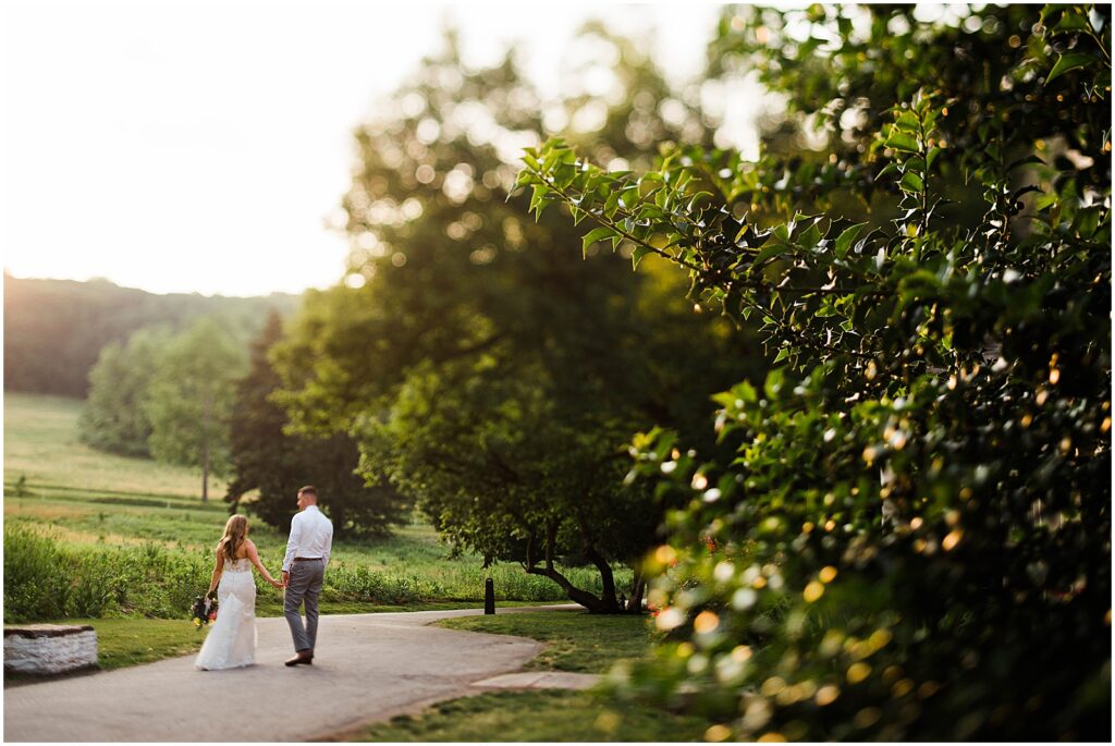 A bride and groom hold hands and walk down a tree-lined path in Valley Forge.