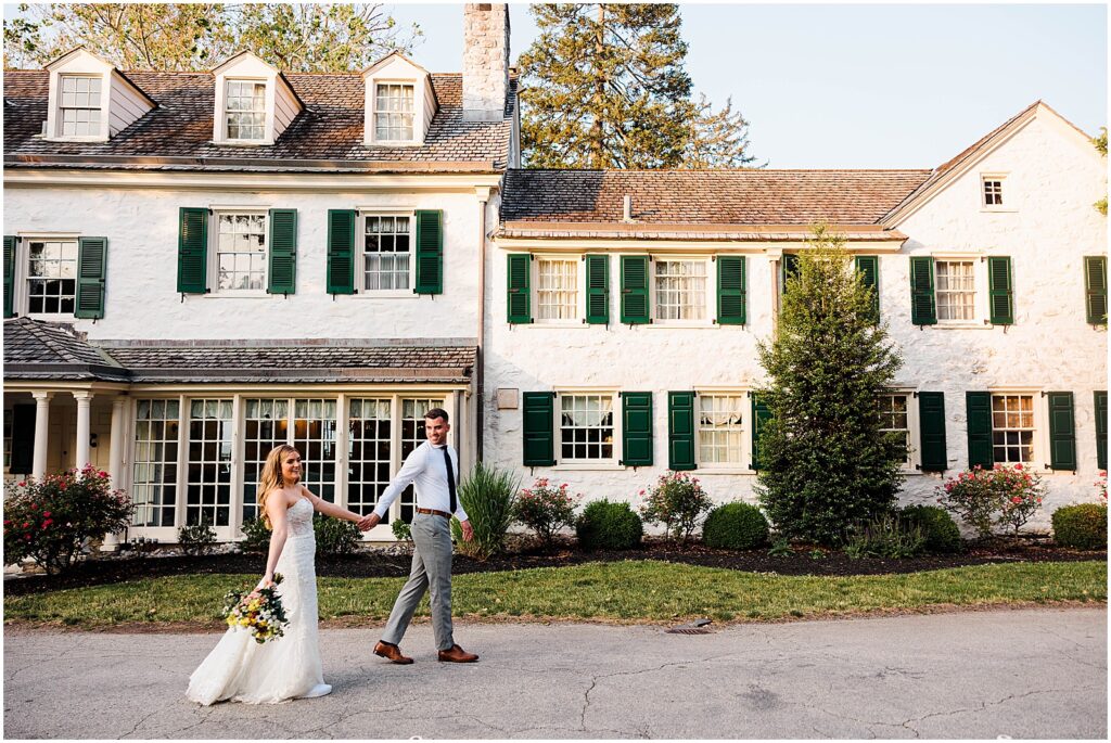 A bride and groom hold hands and look at a New Jersey wedding photographer as they walk in front of the house at Philander Chase Knox Estate.