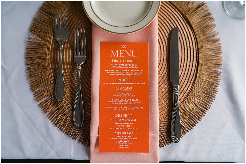 An orange menu sits on a pink napkin on top of a rattan place mat.