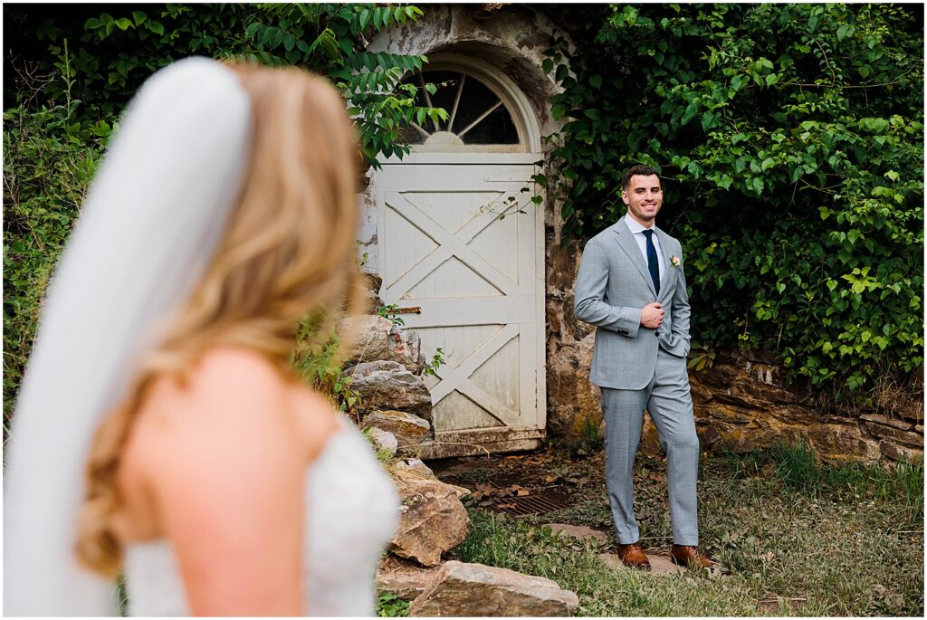 A bride looks across a lawn at a groom posing with his hand in his pocket.