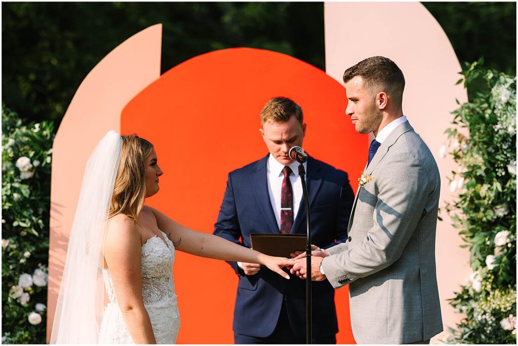 A groom places a wedding ring on a bride in front of a ceremony backdrop.