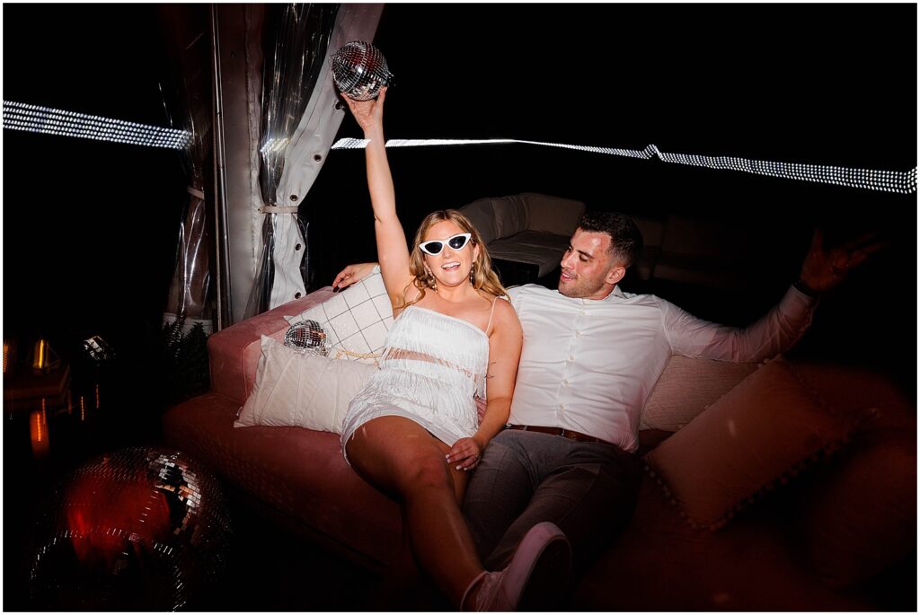 A bride and groom sit on a pink couch and hold up a mirror ball at a disco themed wedding in a direct flash wedding photo.