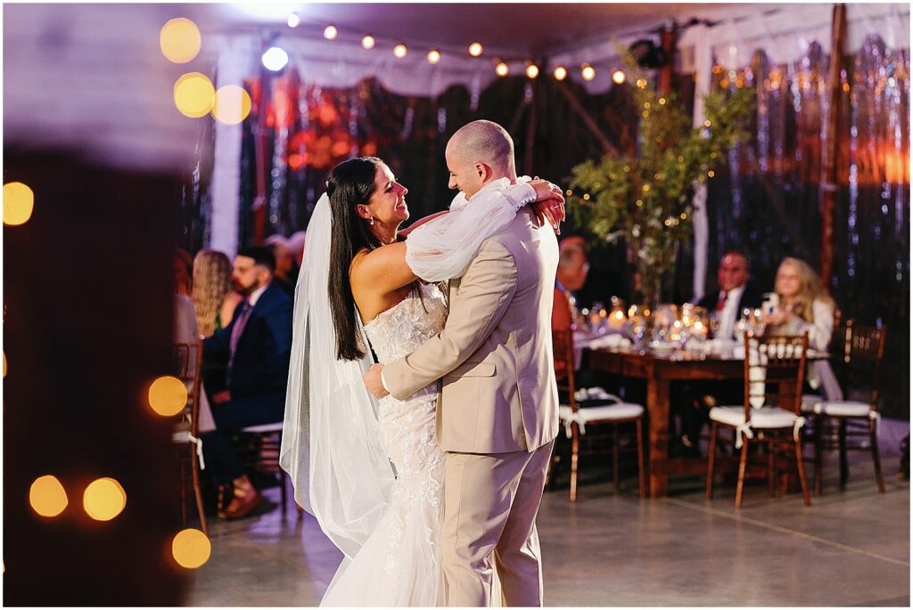 A bride and groom dance while guests watch from reception tables.