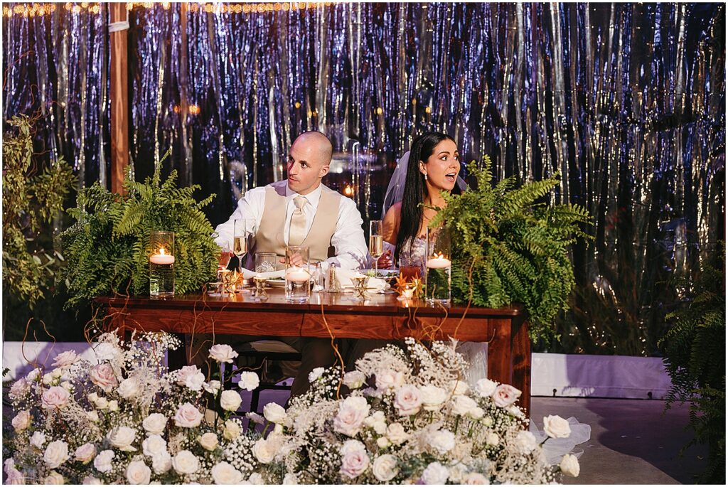 A bride and groom sit at a sweetheart table decorated with ferns.