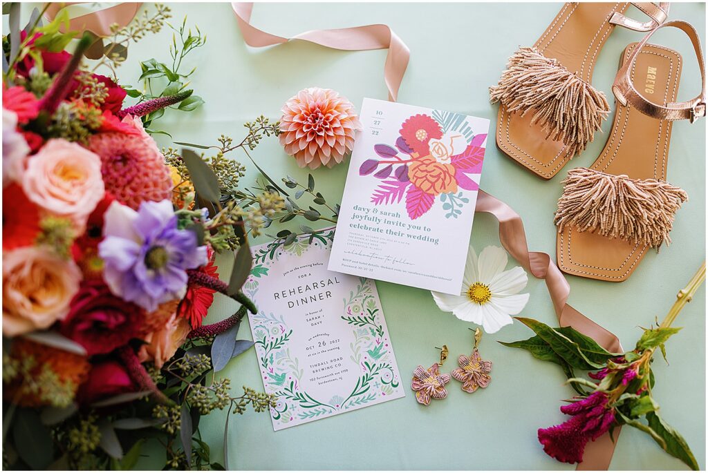 Colorful wedding invitations sit on a table with a bridal bouquet and wedding shoes at a New Jersey wedding.