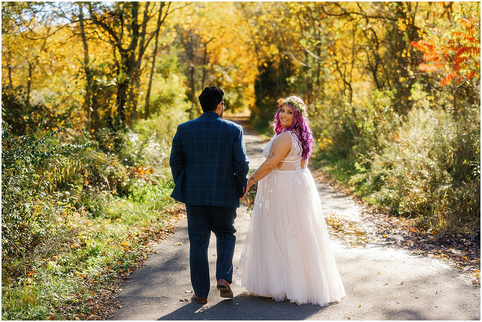 A bride looks over her shoulder as she walks down a path at the Estate at Eagle Lake holding hands with a groom.