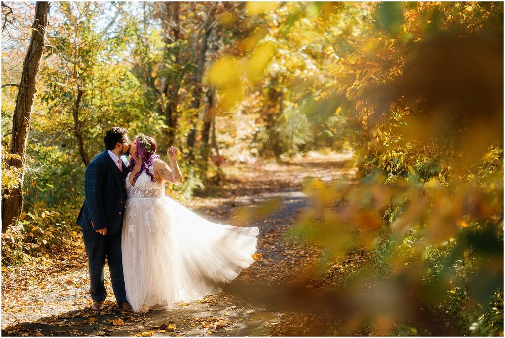 A bride and groom kiss beside yellow-leafed trees at a fall wedding.