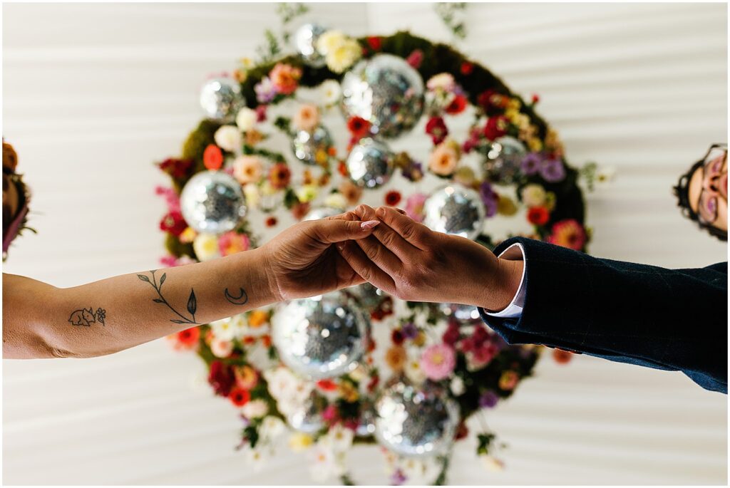 A bride and groom touch hands beneath a wedding installation with bright flowers and disco balls.