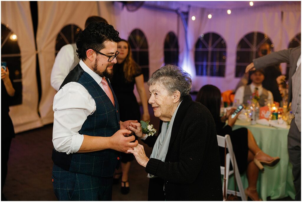 A groom holds an elderly family member's hands beside a wedding reception table.