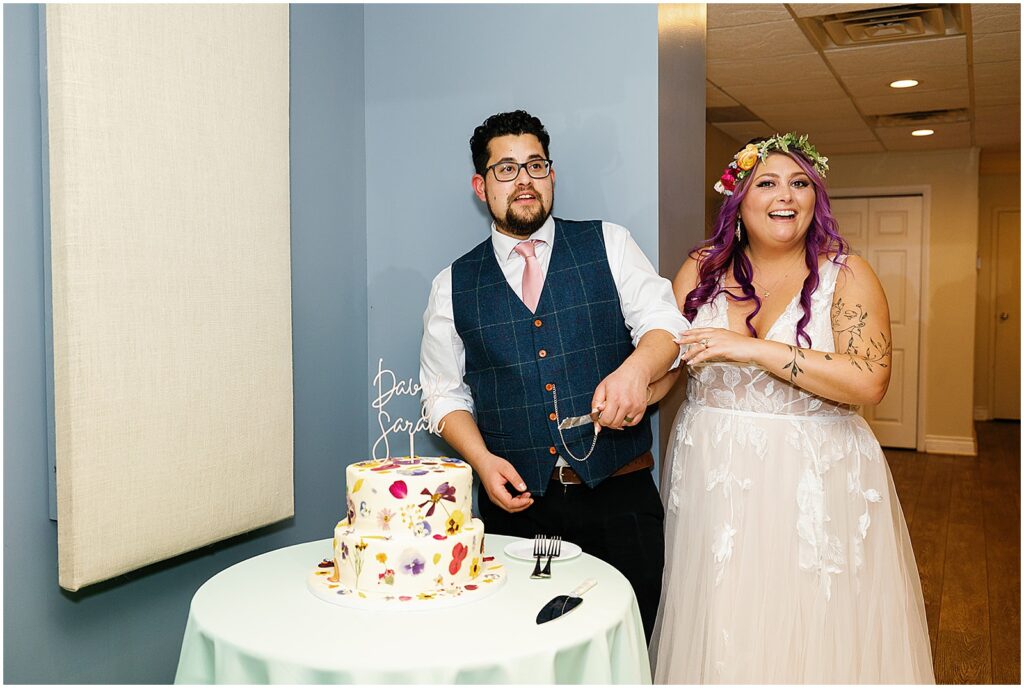 A bride and groom laugh beside a wedding cake decorated with pressed flowers.