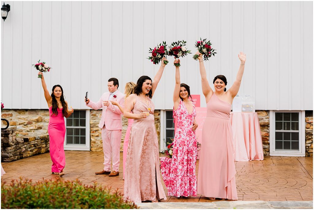 Bridesmaids in mismatched pink bridesmaids dresses wave their bouquets outside the Barn at Stoneybrooke.