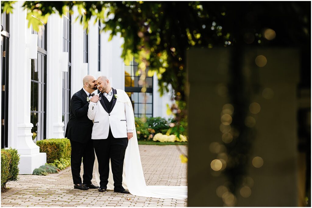 Two grooms wearing black and white wedding details cuddle in a courtyard wedding venue.