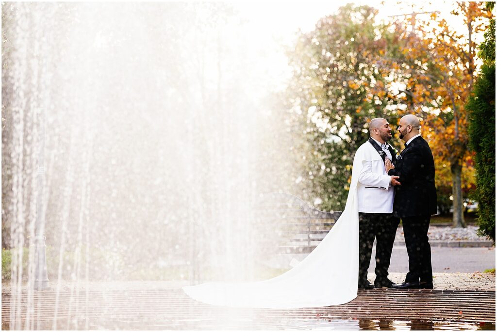 Two grooms pose for candid wedding photos behind a fountain.