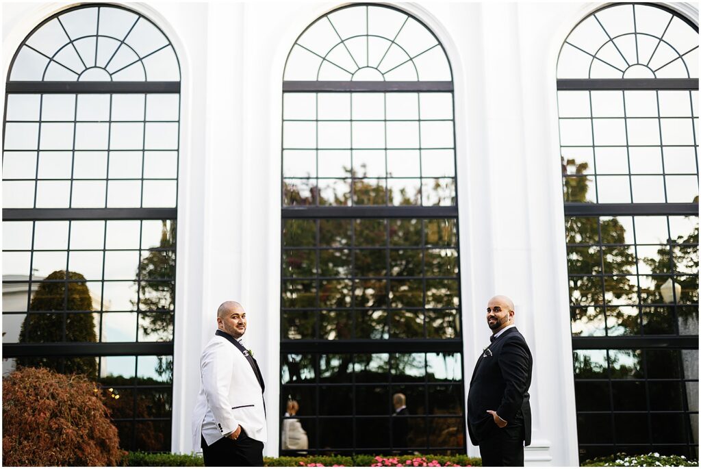 Two grooms pose beside tall windows for editorial wedding photos.