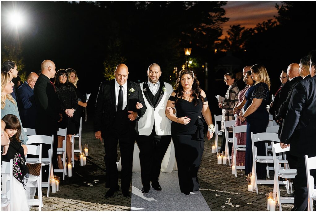 A groom's parents walk them up the aisle for a nighttime wedding ceremony.