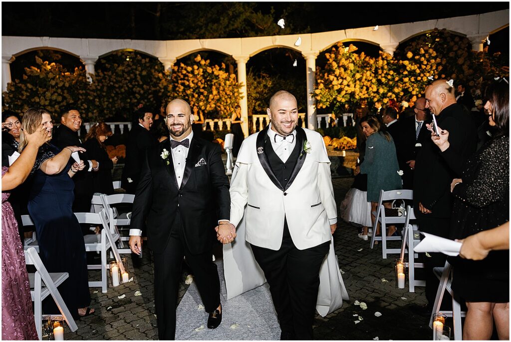 Two grooms hold hands as they walk the recessional at their nighttime wedding ceremony.