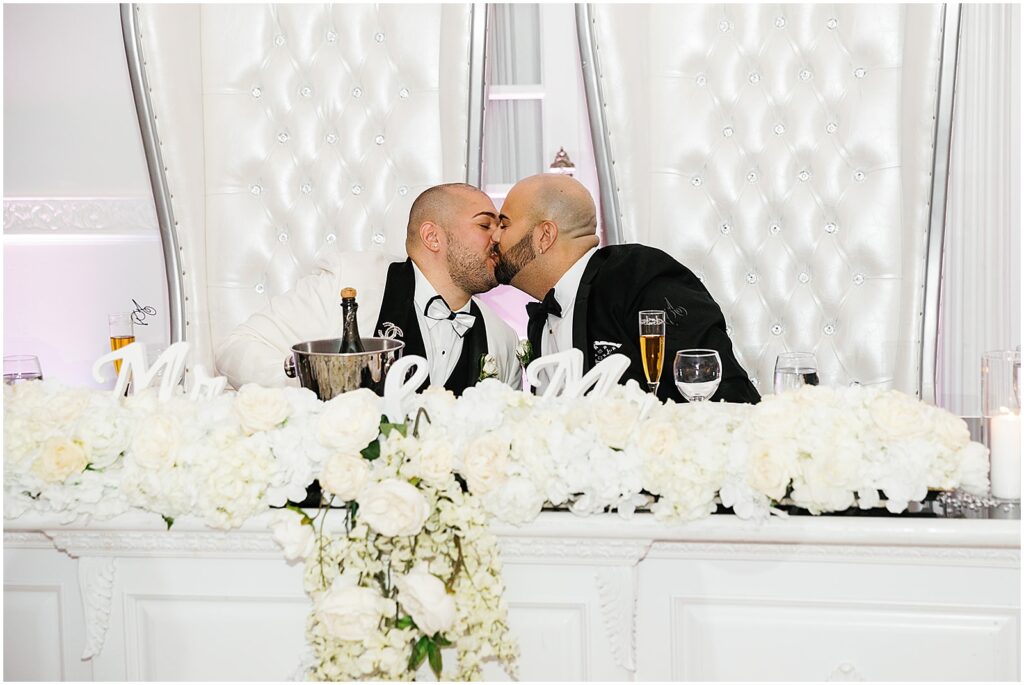 Two grooms kiss at a sweetheart table decorated with white wedding flowers.