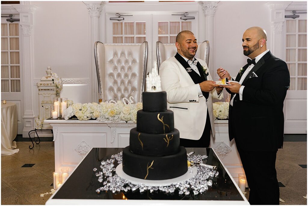 Two grooms stand beside a black wedding cake.