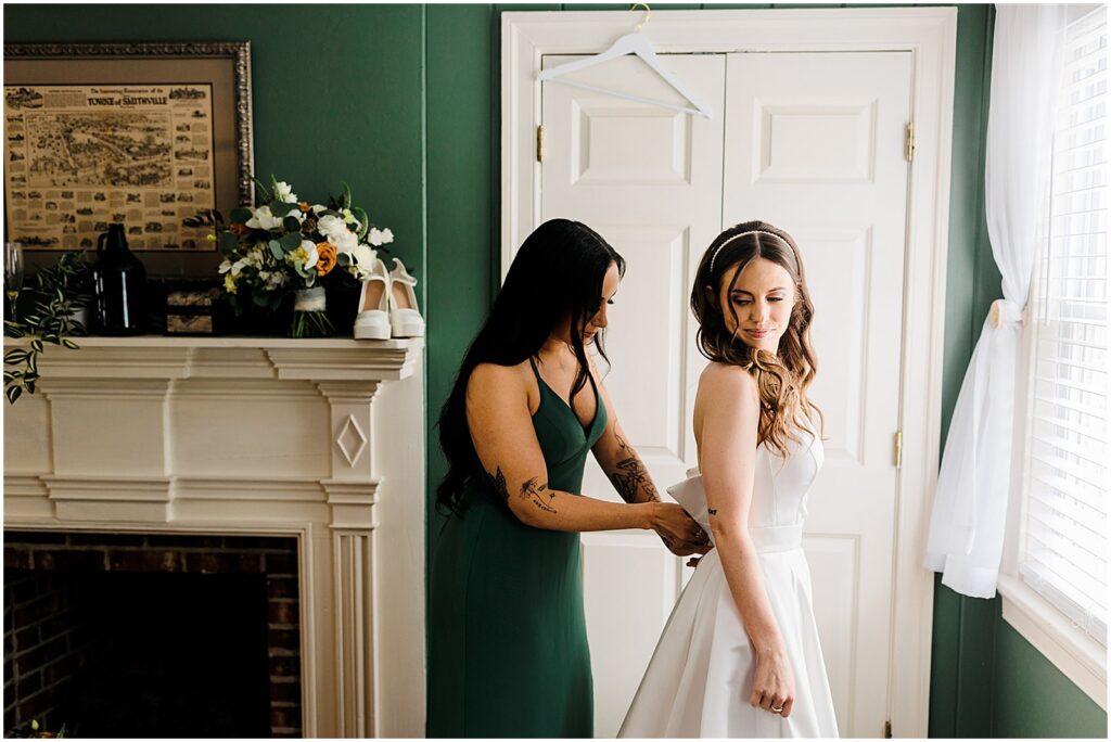 A woman wearing a green bridesmaid dress helps a bride fasten her wedding dress beside a window.