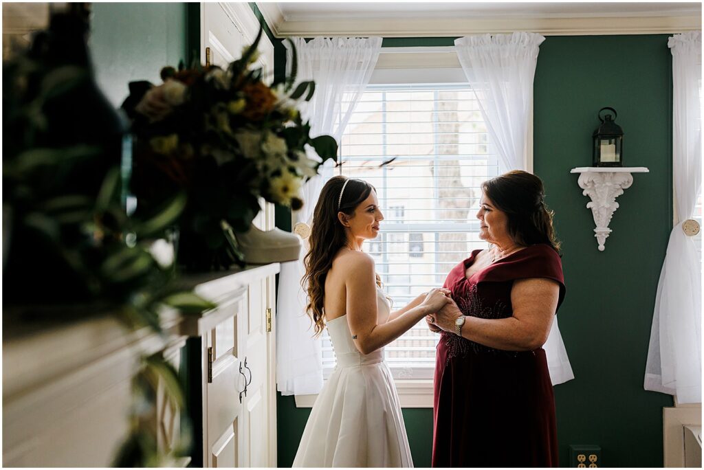 A bride holds hands with her mother in front of a window.