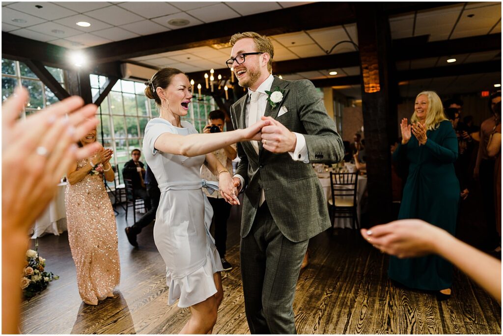 A groom dances with a wedding guest while others clap.