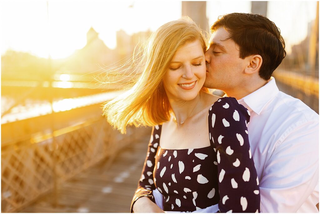 A man kisses a woman's cheek on the Brooklyn Bridge in sunrise Brooklyn engagement photos.