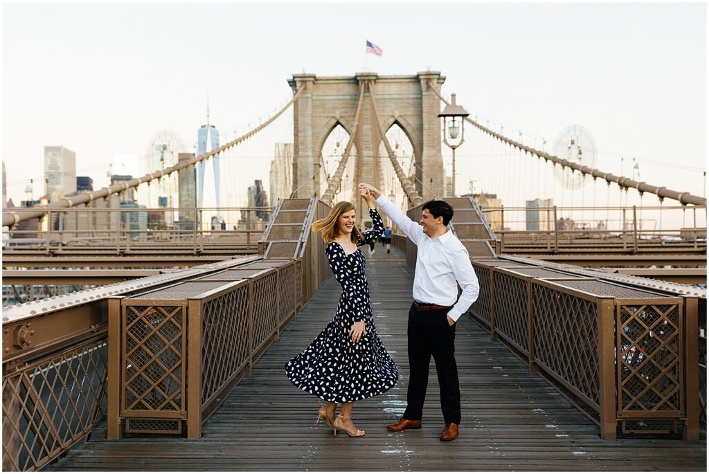 A man twirls a woman on the Brooklyn Bridge for their Brooklyn engagement photos.