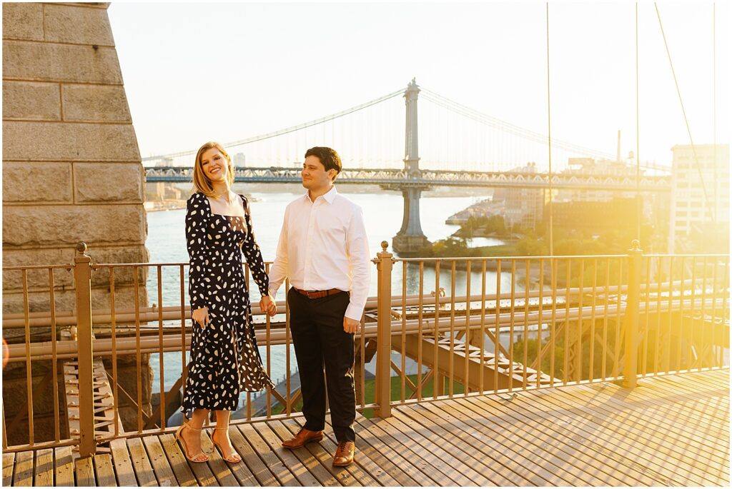 A man and woman laugh on a boardwalk with the Manhattan Bridge behind them.