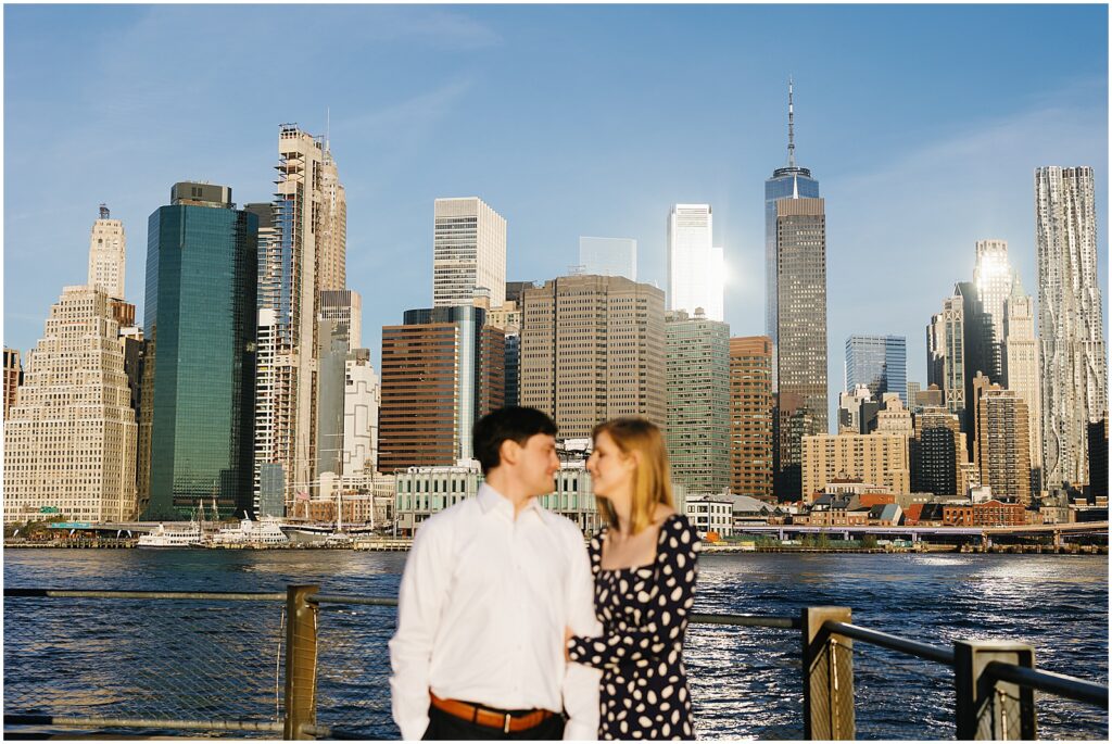 A man and woman pose for an engagement photo at Brooklyn Bridge Park with the Manhattan skyline behind them.