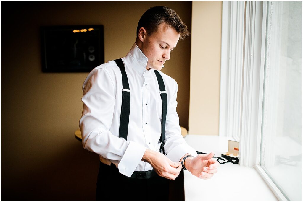 A groom buttons a cufflink beside a window.