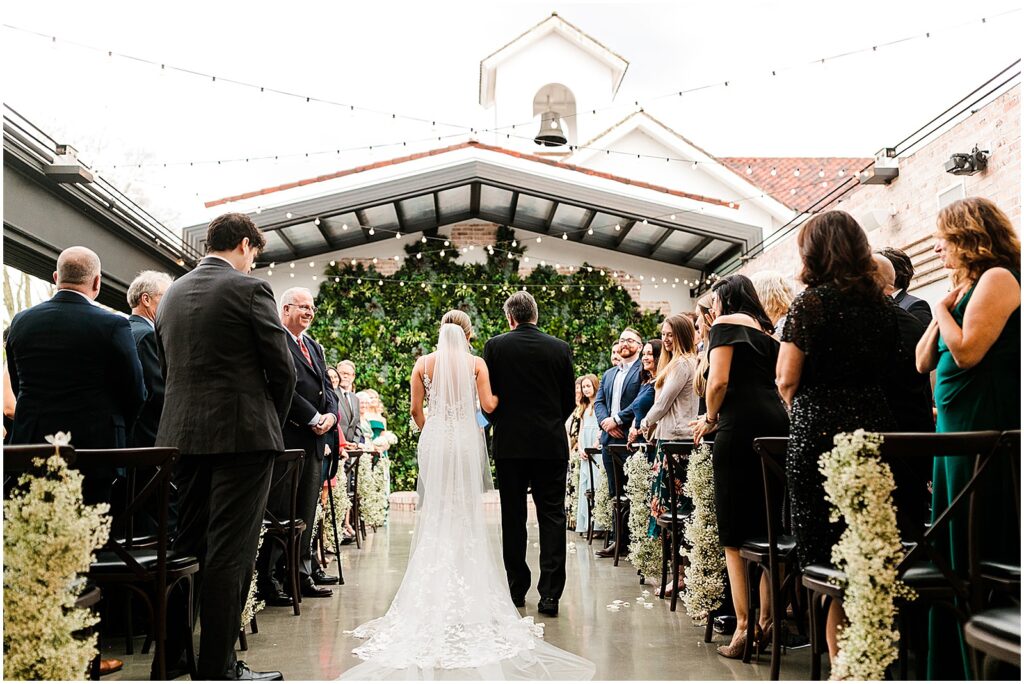 A bride and her father stand at the beginning of an aisle as wedding guests stand.