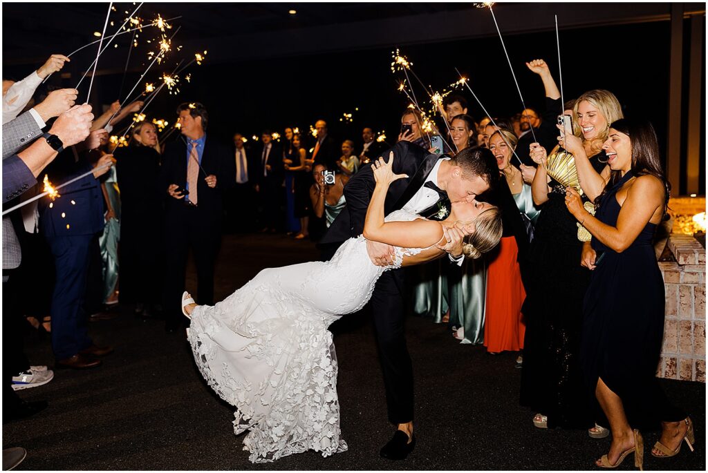 A groom dips a bride at the end of a line of wedding guests holding sparklers.