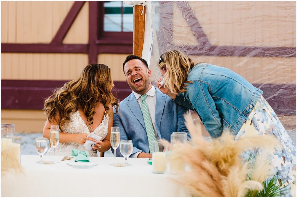 A bridesmaid leans over the sweetheart table and laughs with the bride and groom.