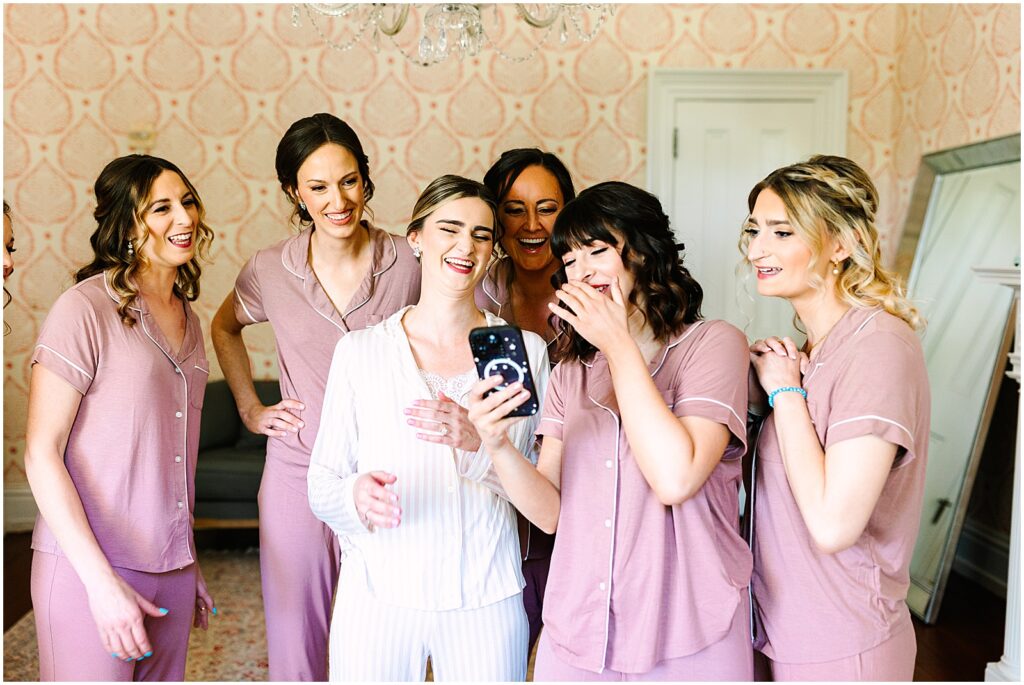 A bride and her bridesmaids laugh at something on a phone.