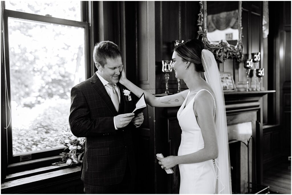 A bride puts her hand on a groom's cheek while he reads his vows in a library at Bellevue Hall.