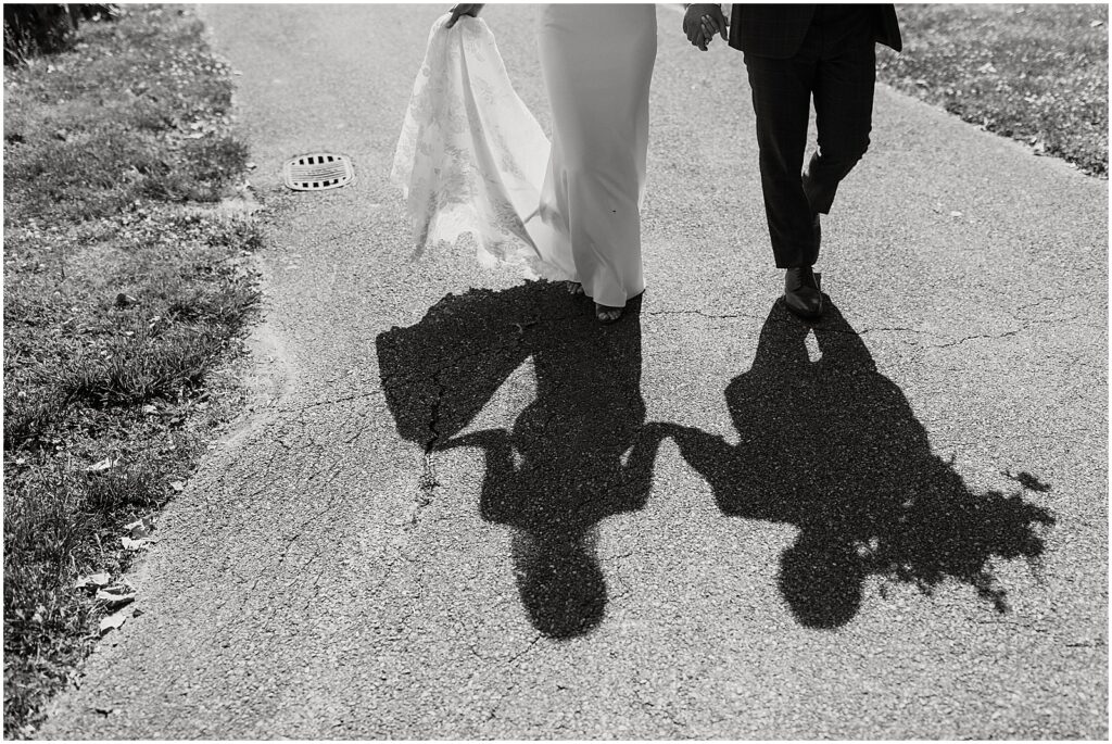A bride and groom cast shadows as they walk down a path at a May wedding.
