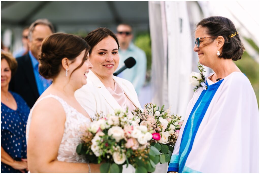 An officiant in a white and blue robe smiles at two brides.