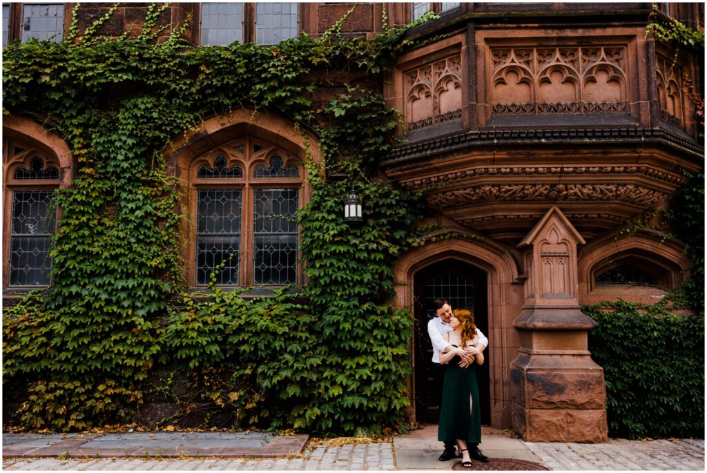 A man and woman pose for creative engagement photos in front of an ivy-covered building.