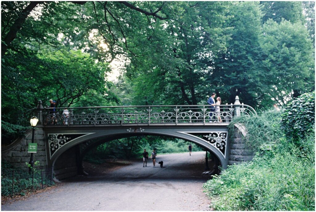 A man and woman stand on a bridge in Central Park in an engagement photo on film.