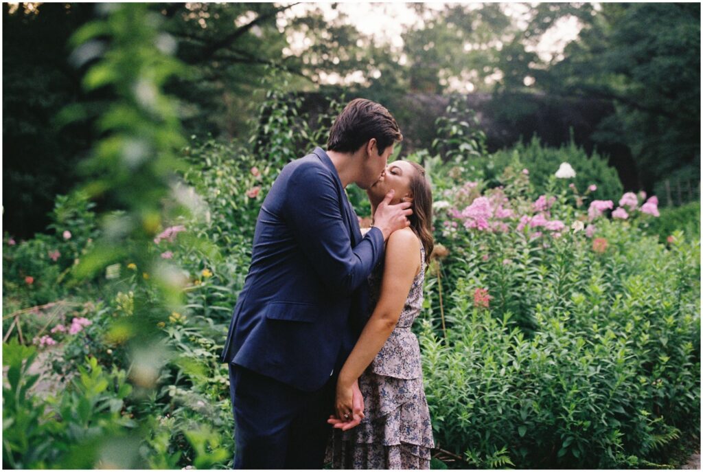 A man and woman kiss in a Central Park garden in New York City engagement photos.