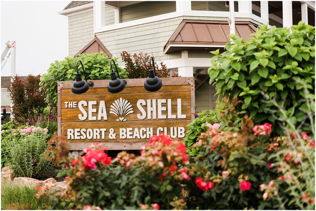 Flowers bloom around a wooden sign for the Sea Shell Resort and Beach Club.